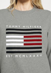Sudadera gris con un diseño de rayas horizontales en negro, blanco, rojo y rojo oscuro. El texto dice "TOMMY HILFIGER" y "EST. MCMLXXXV."