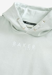 Heleroheline kapuutsiga dressipluus, millel on kängurutasku; valge tikandiga bränding ja metallist paelasilmad; pehme ja sile tekstuur.