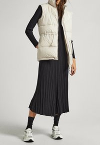 Beżowa, pikowana kamizelka bez rękawów z przednimi kieszeniami, noszona na czarnej, prążkowanej sukience maxi. Uzupełniona czarnymi skarpetkami i białymi trampkami.