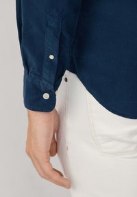 Marineblauwe corduroy shirt met knoopmanchetten en een gebogen zoom, gecombineerd met witte broek. De stof heeft een geribbelde textuur en een gladde afwerking.