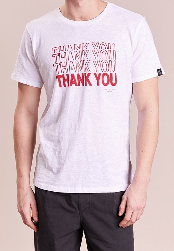 Homme portant un T-shirt blanc avec le texte rouge répétant « MERCI » en différentes intensités de gras, associé à un pantalon sombre.