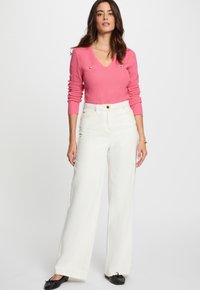 Femme se tenant debout, portant un pull à col en V rose à manches longues, un pantalon large taille haute blanc, des ballerines noires et des bijoux en or.