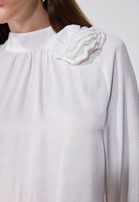 Hvit bluse med høy krage og rynkedetaljer. Har en stor, teksturert stoffblomst som aksent på venstre skulder. Jevn stofftekstur.
