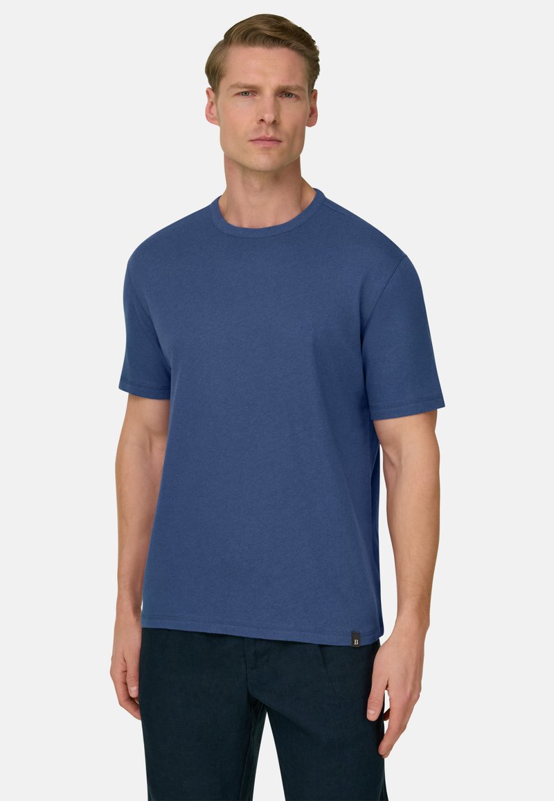 Homme adulte à la peau claire et aux cheveux blonds courts, portant un t-shirt bleu uni à manches courtes et un pantalon foncé, debout devant un fond uni.