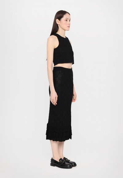 Fekete kötött cropped top és testhezálló midi szoknya fodros szegéllyel; bonyolult textúra és minta; fekete slip-on cipőkkel párosítva, amelyek fémes díszítéssel rendelkeznek.