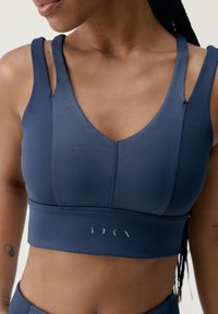 Jūros mėlynos sportinės liemenėlės V formos iškirptė, su platesnėmis petnešėlėmis, siuvinėtomis siūlėmis ir logotipu apatinėje juostoje. Lygus paviršius.