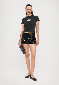 Top a maniche corte nera fitted con un taglio unico, abbinato a pantaloni corti neri lucidi e una borsa a pochette blu con texture. A completare il look, semplici ballerine nere.