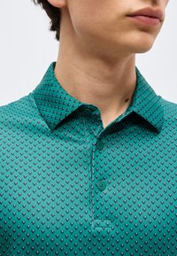 Κοντινό πλάνο ενός ατόμου που φοράει πράσινο πουκάμισο με γιακά, με μικρό σκούρο καρδιακό μοτίβο και τρία πράσινα κουμπιά στην πατιλέτα.