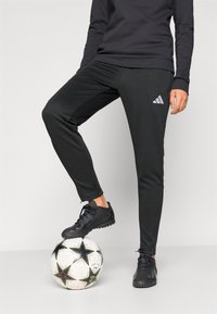 Μαύρο αθλητικό παντελόνι με εφαρμοστή γραμμή, με λογότυπο της Adidas. Το άτομο στέκεται πάνω σε μια ποδοσφαιρική μπάλα με μαύρο και λευκό μοτίβο αστέρων.