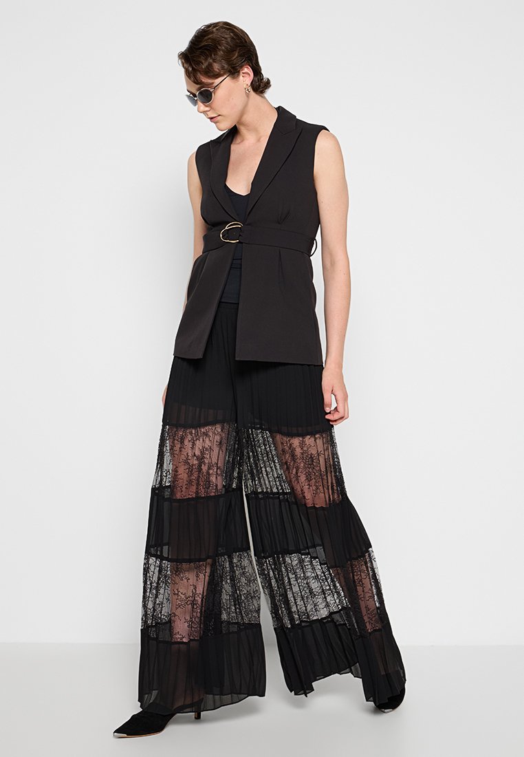 Gilet sans manches noir avec un détail de ceinture, associé à un pantalon noir à jambes larges comportant des panneaux en dentelle transparente et des plis. Stylé avec des lunettes de soleil.