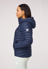 Marineblauwe gewatteerde jas met capuchon, voorzien van een gequilte uitstraling, zijzakken en een klein logopatch op de linkermouw. Glad textuur.
