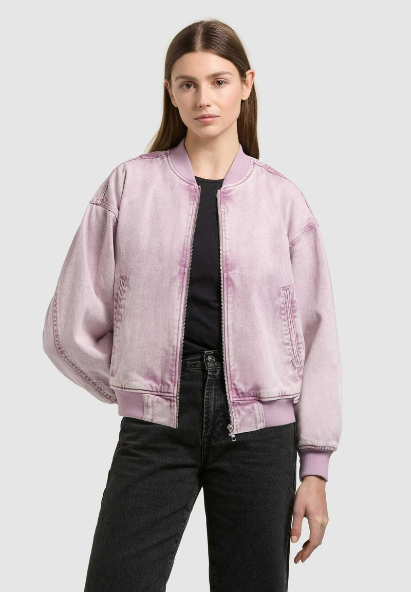 Jeune femme aux cheveux bruns et raides portant une veste bomber violet clair sur un haut noir et un jean noir, debout devant un fond gris.