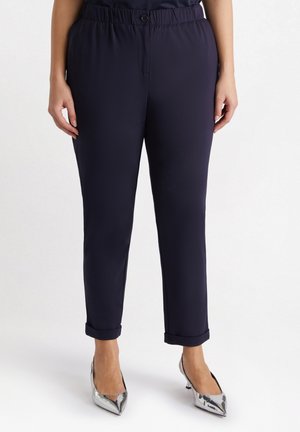 Donkerblauwe, rekbare broek met een zachte textuur; heeft een knoop bij de tailleband en opgerolde pijpen, gecombineerd met puntige zilveren hakken.