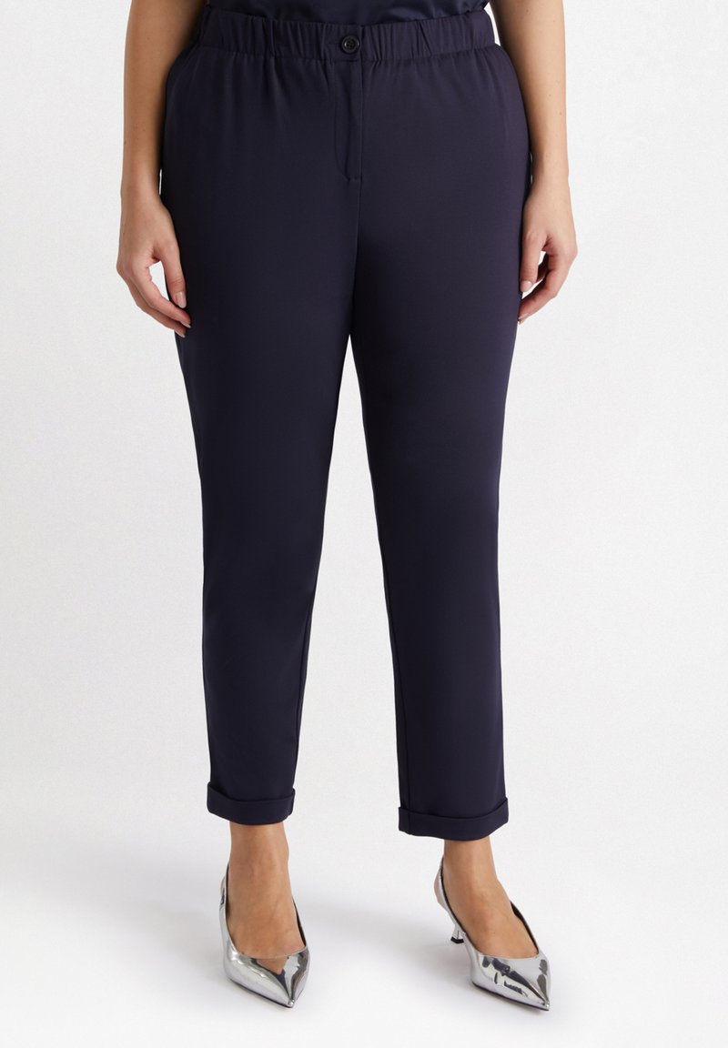 Pantalon extensible bleu marine avec une texture lisse ; comprend un bouton à la taille et des ourlets roulés, associé à des talons argentés pointus.