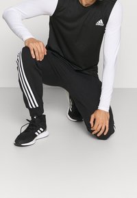 Top sportivo nero senza maniche con maniche lunghe bianche, abbinato a pantaloni neri con tre strisce bianche. Sneakers nere con dettagli bianchi.