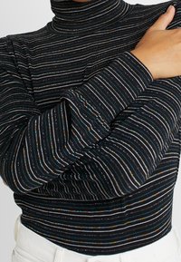 Haut à col roulé noir avec des rayures horizontales multicolores ornées d'accents métalliques. Le tissu présente une texture côtelée et une coupe ajustée.