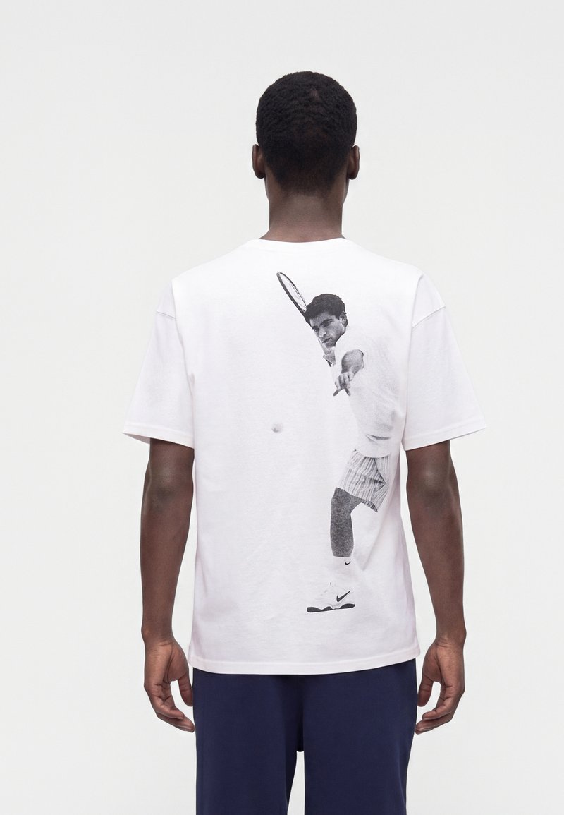 Mand iført en hvid t-shirt med et sort-hvidt print af en tennisspiller, der slår en bold på ryggen, kombineret med mørkeblå bukser.
