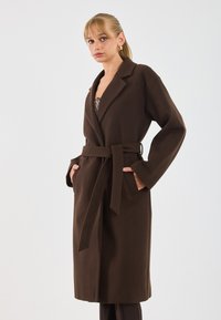 Donna con capelli biondi che indossa orecchini a cerchio d'oro e un lungo cappotto marrone con cintura, con le mani in tasca, su uno sfondo neutro.