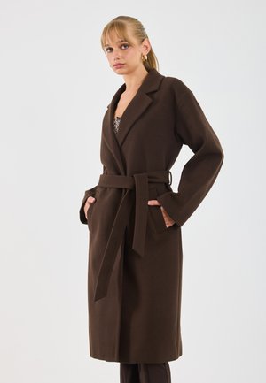 Donna con capelli biondi che indossa orecchini a cerchio d'oro e un lungo cappotto marrone con cintura, con le mani in tasca, su uno sfondo neutro.