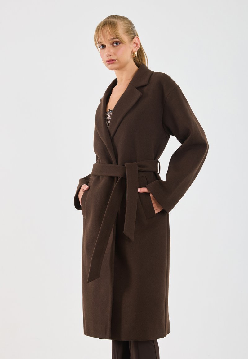 Donna con capelli biondi che indossa orecchini a cerchio d'oro e un lungo cappotto marrone con cintura, con le mani in tasca, su uno sfondo neutro.
