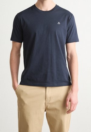 Mężczyzna ubrany w granatową koszulkę z krótkim rękawem z małym białym logo na piersi oraz beżowe spodnie, stojący z jedną ręką w kieszeni.