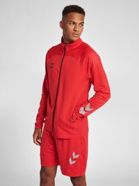 Rød atletisk jakke med lynlås foran og høj krave, parret med matchende shorts. Har struktureret stof og sølvdetaljer ved ærmerne og shortsene.