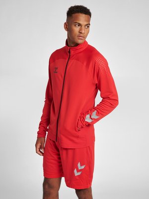 Rød atletisk jakke med lynlås foran og høj krave, parret med matchende shorts. Har struktureret stof og sølvdetaljer ved ærmerne og shortsene.