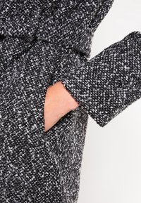 Abrigo texturizado en blanco y negro con un bolsillo visible, que presenta una cintura ajustada y un puño ancho en la manga, mostrando un patrón moteado.