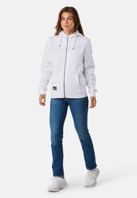 Weißer Kapuzenpullover mit Reißverschluss und „SEASIDE“ auf der Vorderseite, Fronttaschen und Kordelzugkapuze, kombiniert mit blauen Jeans und weißen Sneakers.