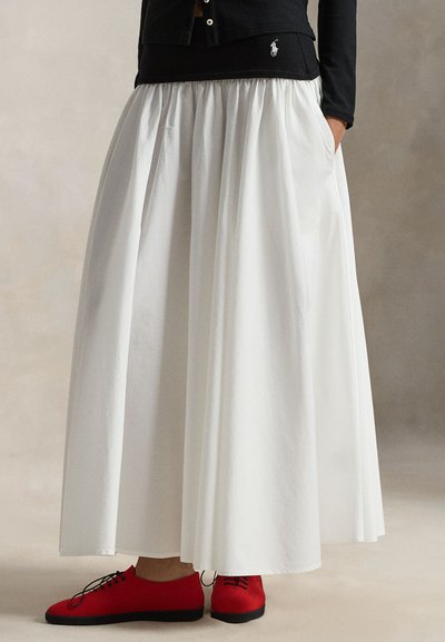 Jupe blanche longue jusqu'aux pieds avec une taille froncée, associée à des chaussures rouges. La jupe présente une texture lisse et une silhouette fluide.