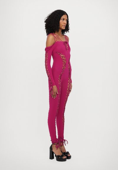 Salopetă fucsia, adaptată pe corp, cu design off-shoulder, detalii de șiret pe laterale și legături la glezne. Asortată cu pantofi cu platformă negri.