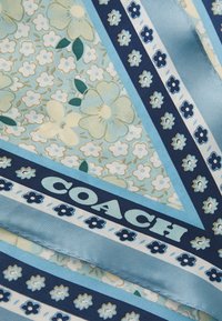Μεταξωτό κασκόλ με ανθισμένα σχέδια σε ανοιχτό μπλε και ναυτικό, με διαγώνιες ρίγες και λογότυπο "COACH" σε έντονη γραφή κατά μήκος της άκρης.