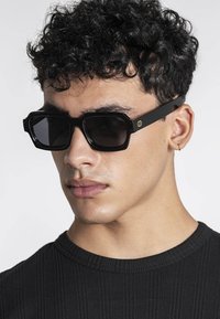 Joven con cabello rizado que lleva gafas de sol negras rectangulares y un pequeño pendiente de aro dorado, vistiendo una camisa negra acanalada.