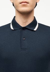 Jeune personne portant un polo bleu marine avec un bord blanc sur le col, boutonné en haut.