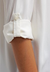 White long-sleeve shirt with a rolled-up cuff featuring a button closure. Smooth, lightweight fabric with a subtle sheen.