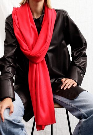 Personne portant une écharpe rouge vif sur une veste en cuir noire et un jean bleu clair, assise avec les mains posées sur les cuisses.