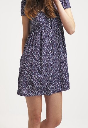 Femme portant une robe courte à boutons sur le devant, avec un motif floral violet et des manches courtes, debout devant un fond clair uni.
