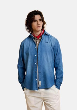 Jonge man met halflang donker haar, gekleed in een blauw spijkerhemd over een beige polo, rode zakdoek om de hals en witte broek, handen in de zakken.