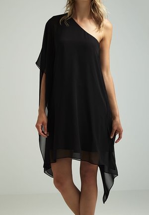 Femme portant une robe en chiffon noire asymétrique à une épaule, avec une coupe ample et fluide et des détails transparents à l'ourlet.