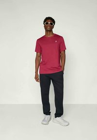 Bordeauxrotes T-Shirt mit kurzen Ärmeln und einem kleinen Logo auf der Brust. Kombiniert mit dunkler Hose und weißen Sneakern, getragen von einem stehenden Model.