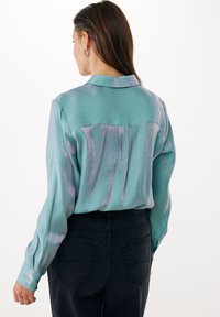 Turquoise satijnen shirt met lange mouwen, een achterpand en subtiele paarse strepen. Gecombineerd met zwarte high-waisted jeans.