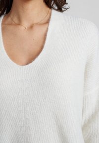 Pull en maille blanc avec un col en V profond, présentant une texture douce et un détail côtelé, associé à un délicat collier en or.