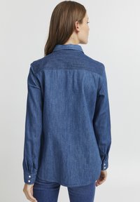 Camicia in denim blu con colletto classico, polsini con bottoni e design dritto sul retro. Presenta una sottile texture e una vestibilità leggermente ampia.