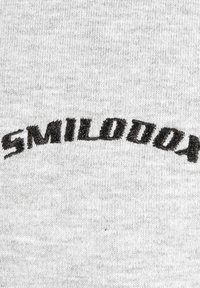 Grauer Stoff mit schwarzem gesticktem Text, der "SMILODON" in einer leicht gebogenen Schrift zeigt und Textur sowie Details hervorhebt.