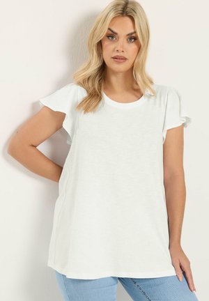 Kobieta z długimi blond włosami, ubrana w prostą białą bluzkę z krótkim rękawem i jasnoniebieskie dżinsy, stojąca z jedną ręką na biodrze.