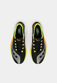 Baskets de sport noires avec des accents jaunes, texture en maille, semelle en caoutchouc et motif de vague distinctif en gris. Lacets en noir et jaune.