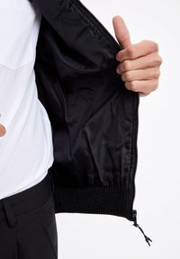 Czarna kurtka bomberka z zamkiem błyskawicznym, wyposażona w gładką podszewkę z satyny oraz ściągacze przy rękawach. Zewnętrzna powierzchnia ma lekką fakturę.