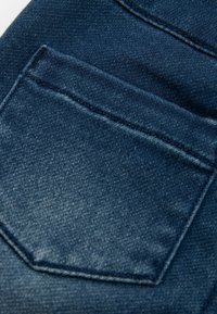 Tessuto in denim blu scuro con una tasca posteriore visibile. La tasca presenta dettagli di cucitura sottili e una texture liscia.