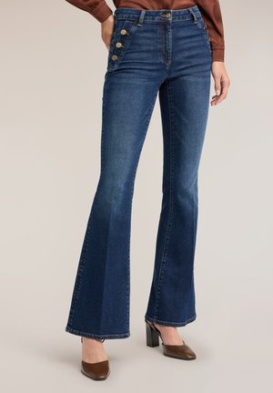 Vrouw draagt donkerblauwe flared jeans met hoge taille en knoopdetails aan de zijkant en bruine hoge hakken, staand tegen een effen achtergrond.