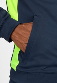 Marineblauwe sportjack met een neon groene accent en een kangaroezak. Glad materiaal met ribgebreide manchetten en zoom.